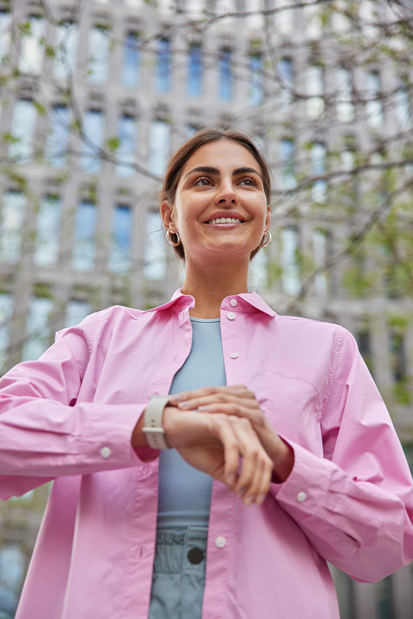 Vertical shot of happy young beautiful woman smiles gently checks time on wrist watch waits for someone comes on meeting wears pink shirt poses against city building. People and lifestyle concept Young beautiful woman happy