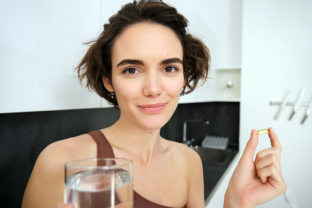 Portrait of sportswoman drinking water, taking vitamins, dietry supplements for healthy skin, having omega-3 pill, standing in her kitchen in workout clothing Oral Treatment in patient with cancer