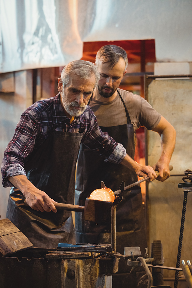 Team of glassblower forming and shaping a molten glass at glassblowing factory Father and son - Happy